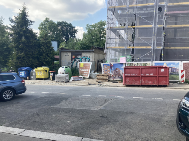 Zwei Autos neben einem Gebäude in Bau neben der Straße mit verstreuten Mülltonnen, Brettern und anderen Gegenständen, Bäumen und einem bewölkten Himmel im Hintergrund.