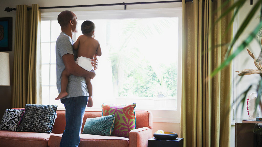 Ein Mann trägt ein Kind vor einem Fenster mit Vorhängen, einer Couch und mehreren Kissen.