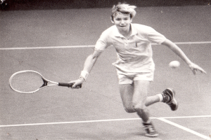 Ein junger Mann in einem weißen Tennis-Outfit schlägt mit einem Schläger auf einen Ball auf einem Tennisplatz und sieht entschlossen aus.