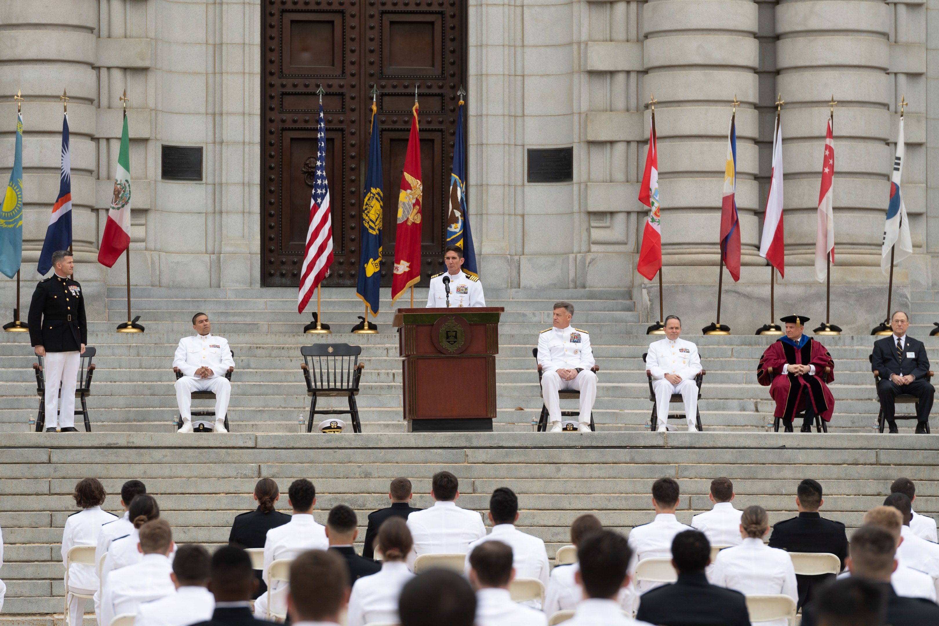 Eine Gruppe von Menschen in weißen Uniformen sitzt auf einer Treppe mit einem Podium, das von Fahnen flankiert ist, vor einem großen Gebäude mit einer Tür.
