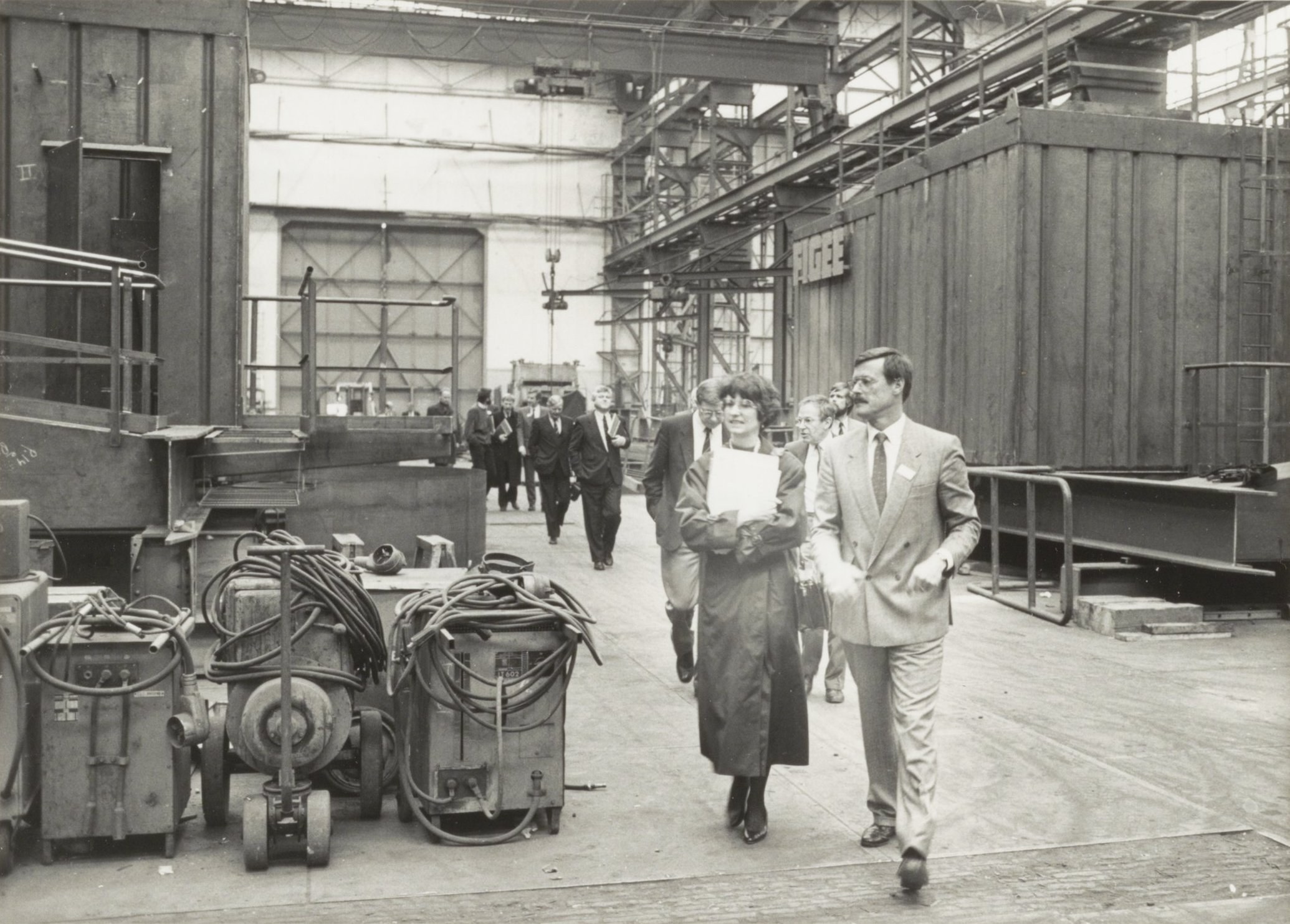 Gruppe von Menschen in Anzügen, die durch eine industrielle Fabrik mit Maschinen auf der linken Seite und einer Wand im Hintergrund gehen.