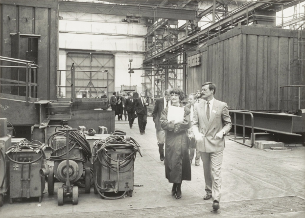 Gruppe von Menschen in Anzügen, die durch eine industrielle Fabrik mit Maschinen auf der linken Seite und einer Wand im Hintergrund gehen.