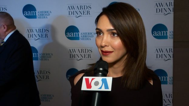Emmy Rossum in einem schwarzen Kleid mit Mikrofon, mit einem Mann links von ihr und einem Banner mit der Aufschrift "Aam Awards Dinner" im Hintergrund.