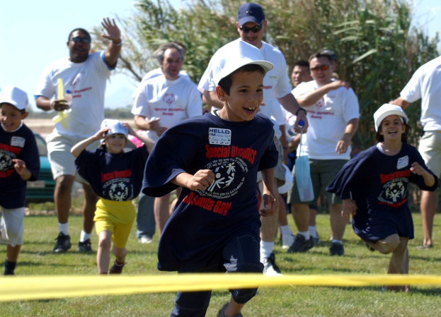 Kinder bei einem Lauf auf einer grünen Wiese mit einem gelben Band im Vordergrund, einige tragen Mützen und Schutzbrillen, andere halten Gegenstände, vor einem klaren blauen Himmel mit Bäumen im Hintergrund.