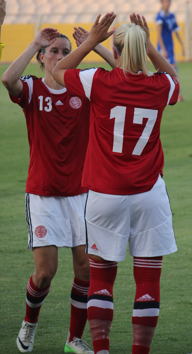 Zwei Frauen in roten T-Shirts und weißen Shorts klatschen auf einem Fußballfeld, mit einer Person in einem blauen Kleid und einem gelben Board im Hintergrund.