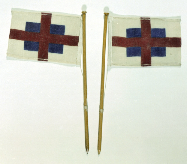 Zwei kleine Flaggen mit Holzstielen, die rote und blaue Kreuze aufweisen, auf einer weißen Oberfläche plaziert.