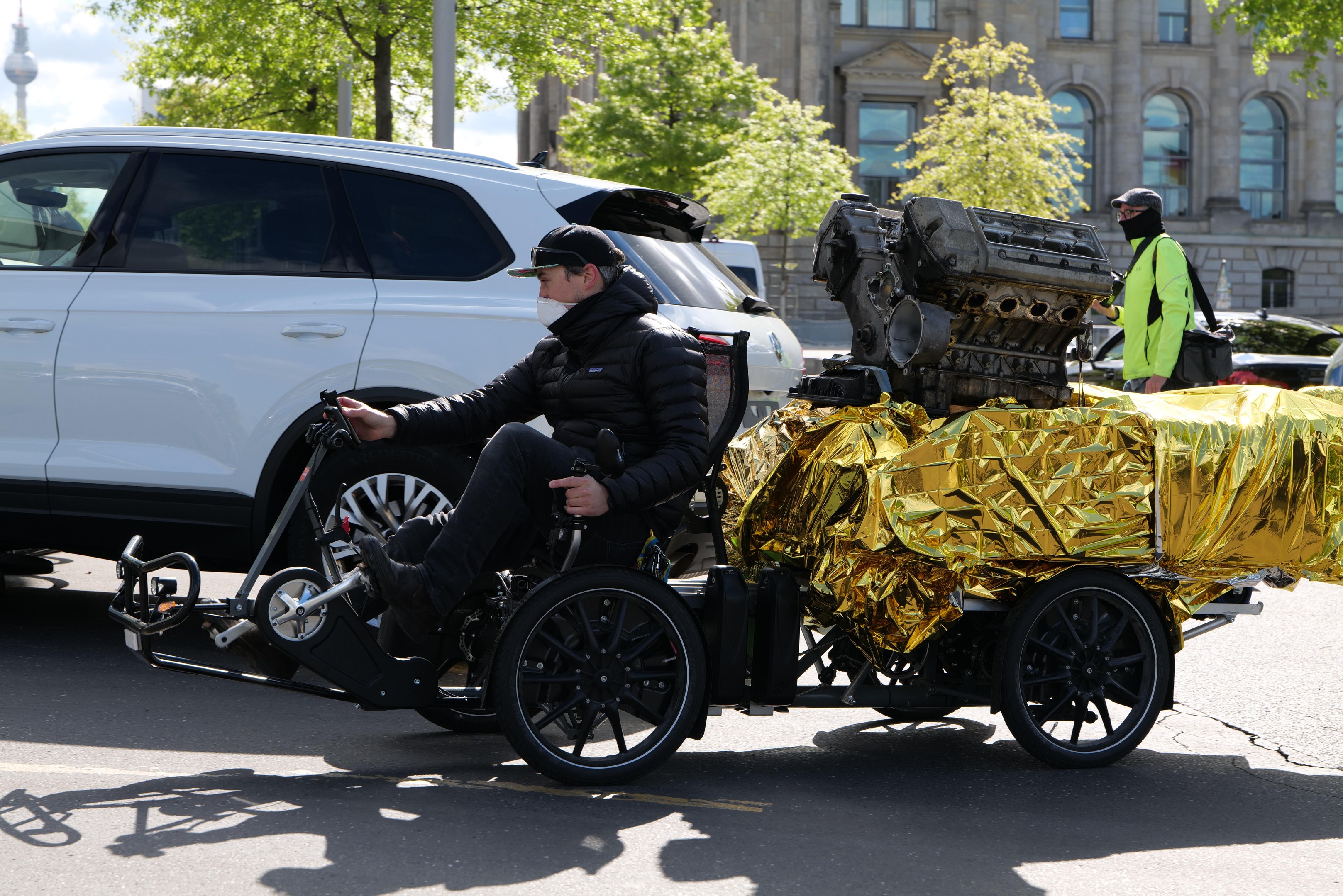 Ein Mann im Rollstuhl mit einem großen Motor daran, umgeben von Fahrzeugen auf einer Straße mit Bäumen, Gebäuden und einem klaren blauen Himmel im Hintergrund, trägt eine schwarze Jacke, eine Mütze und hält ein Objekt.