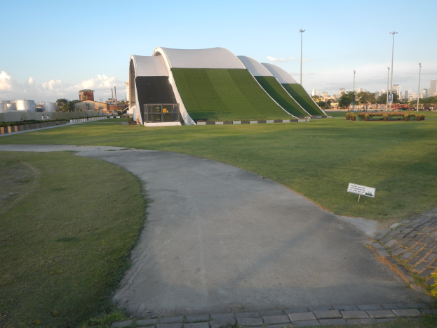Großes Gebäude mit Grünfläche in einem Park mit Rasen, Weg, Informationsschild, Pflanzen, Bäumen, Pfählen, fernen Gebäuden und einem bewölkten Himmel.