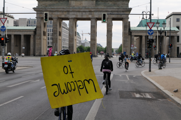 Eine Gruppe von Radfahrern fährt in Helmen eine Straße vor dem Brandenburger Tor in Berlin, Deutschland, entlang, wobei einer ein gelbes Schild hält, Lichtmasten, Verkehrszeichen, Gebäude, Bäume und einen klaren blauen Himmel im Hintergrund.