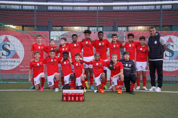 Gruppe junger Männer in roten T-Shirts und schwarzen Shorts auf einem Fußballfeld mit einer Kiste roter Gegenstände davor, umgeben von Bannern mit der Aufschrift "FC Red Black Eglaute - Champions League" und Stadion-Elementen im Hintergrund.