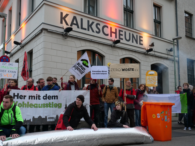 Eine Gruppe von Menschen mit Schildern und Plakaten vor einem Gebäude während einer Demonstration in Deutschland, mit zwei Personen auf einem Objekt im Vordergrund und einem Müllcontainer auf der rechten Seite.
