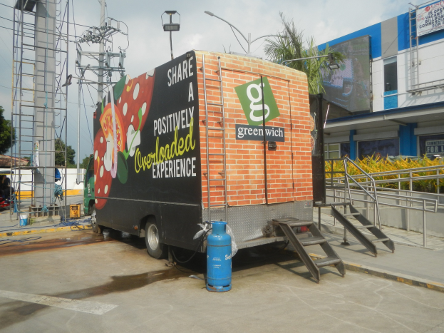Lkw vor einem Gebäude mit einer Tafel, auf der "Teilen Sie eine positiv überladene Erfahrung" steht, mit Stufen, einem Zylinder auf dem Boden, Geländern, elektrischen und Lichtmasten, Bäumen und einem bewölkten Himmel.