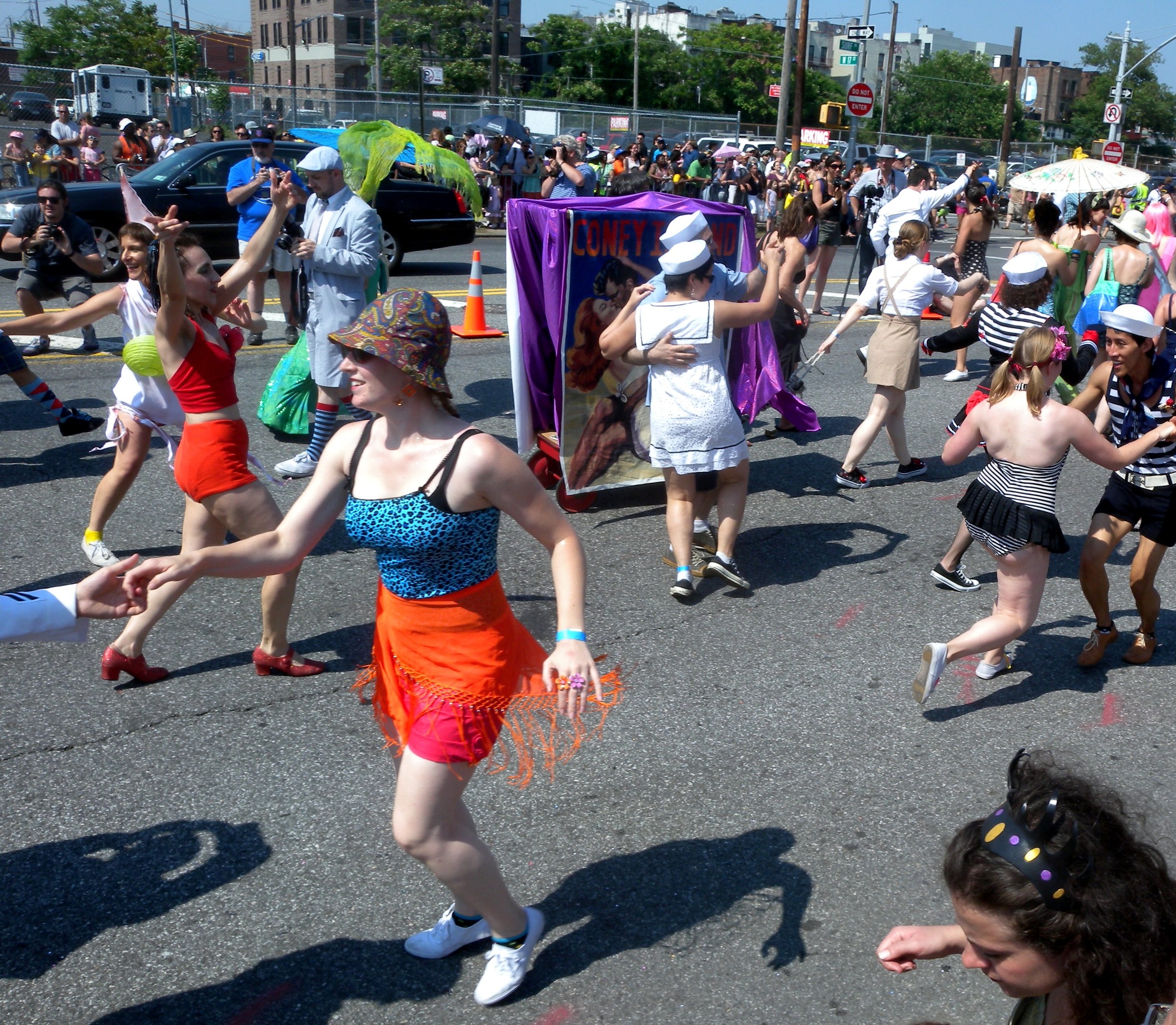 Eine Gruppe von Menschen marschiert auf einer Straße, einige tragen Mützen und halten Schirme, andere tanzen, mit einem Banner, Verkehrskegeln, Fahrzeugen, Laternen, Schildern, Bäumen, Gebäuden und einem klaren blauen Himmel im Hintergrund.