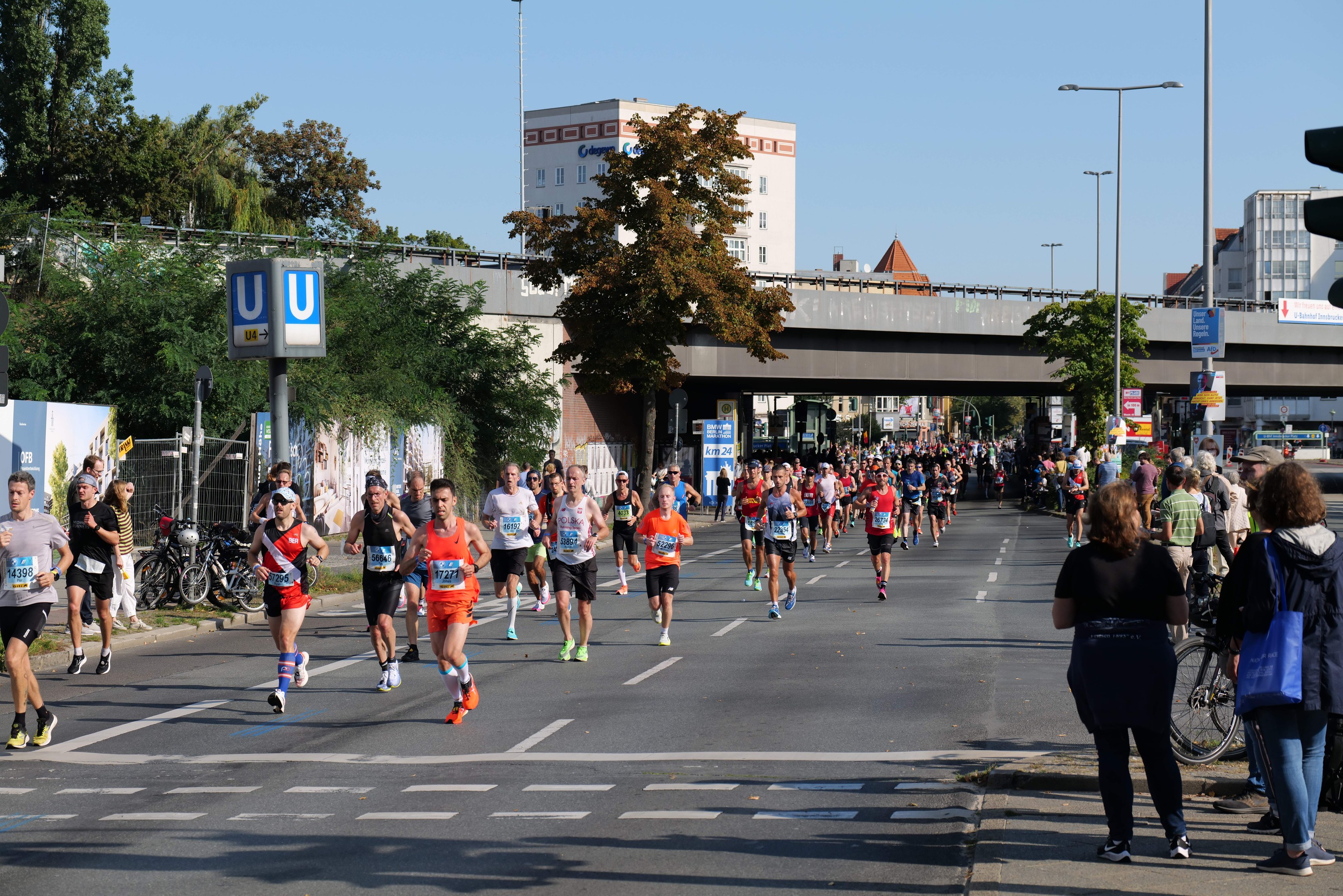 Gruppe von Menschen, die bei einem Marathon auf einer von Bäumen gesäumten Straße laufen, mit Laternen, Schildern, Fahrrädern, einem Zaun, Gras, einer Brücke, Gebäuden und einem klaren blauen Himmel.