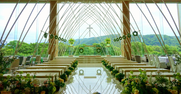 Eine Hochzeitszeremonie in einem großen, geschmückten Saal mit Tischen in Halbkreisform, umgeben von Blumenschmuck, Bäumen, Hügeln und einem klaren blauen Himmel.