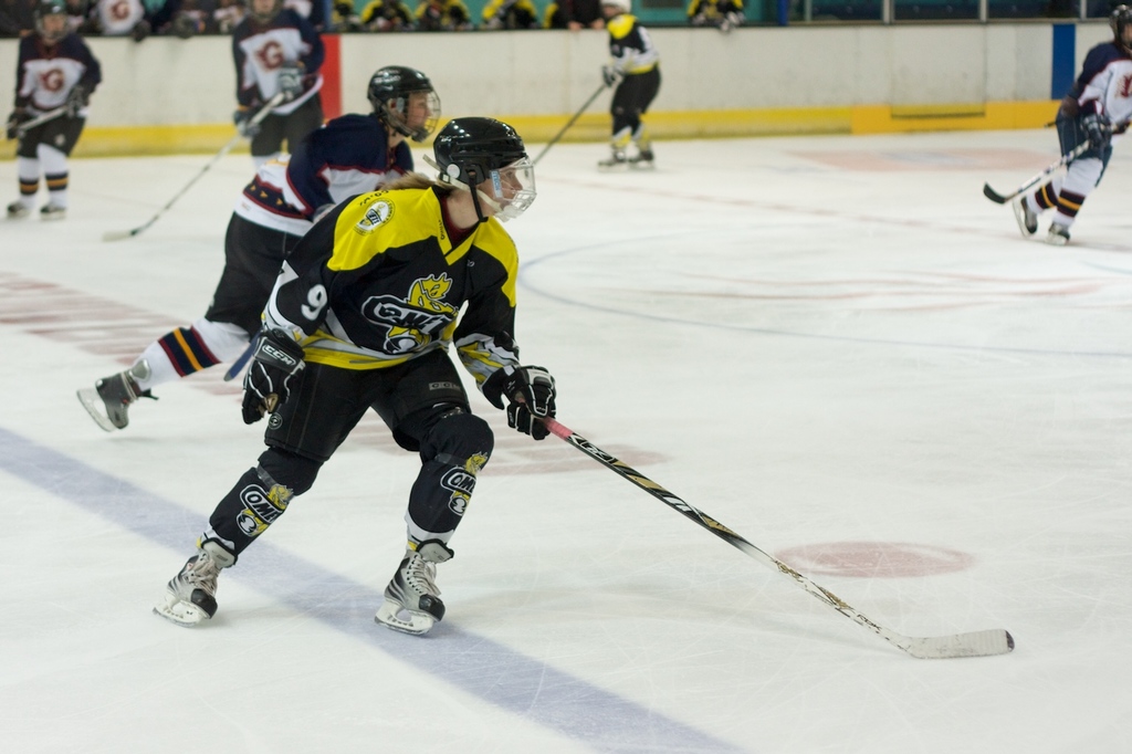 Menschen spielen Eisstockschießen mit Eishockeyschlägern und Helmen, mit anderen und einer Wand im Hintergrund.
