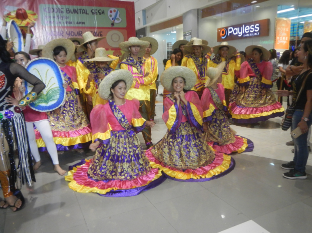 Eine Gruppe von Menschen in farbenfrohen Kleidern und Hüten, die in einem Einkaufszentrum tanzen, während andere in der Nähe mit Musikinstrumenten und einem Banner stehen, unter Deckenleuchten mit Texttafeln im Hintergrund.