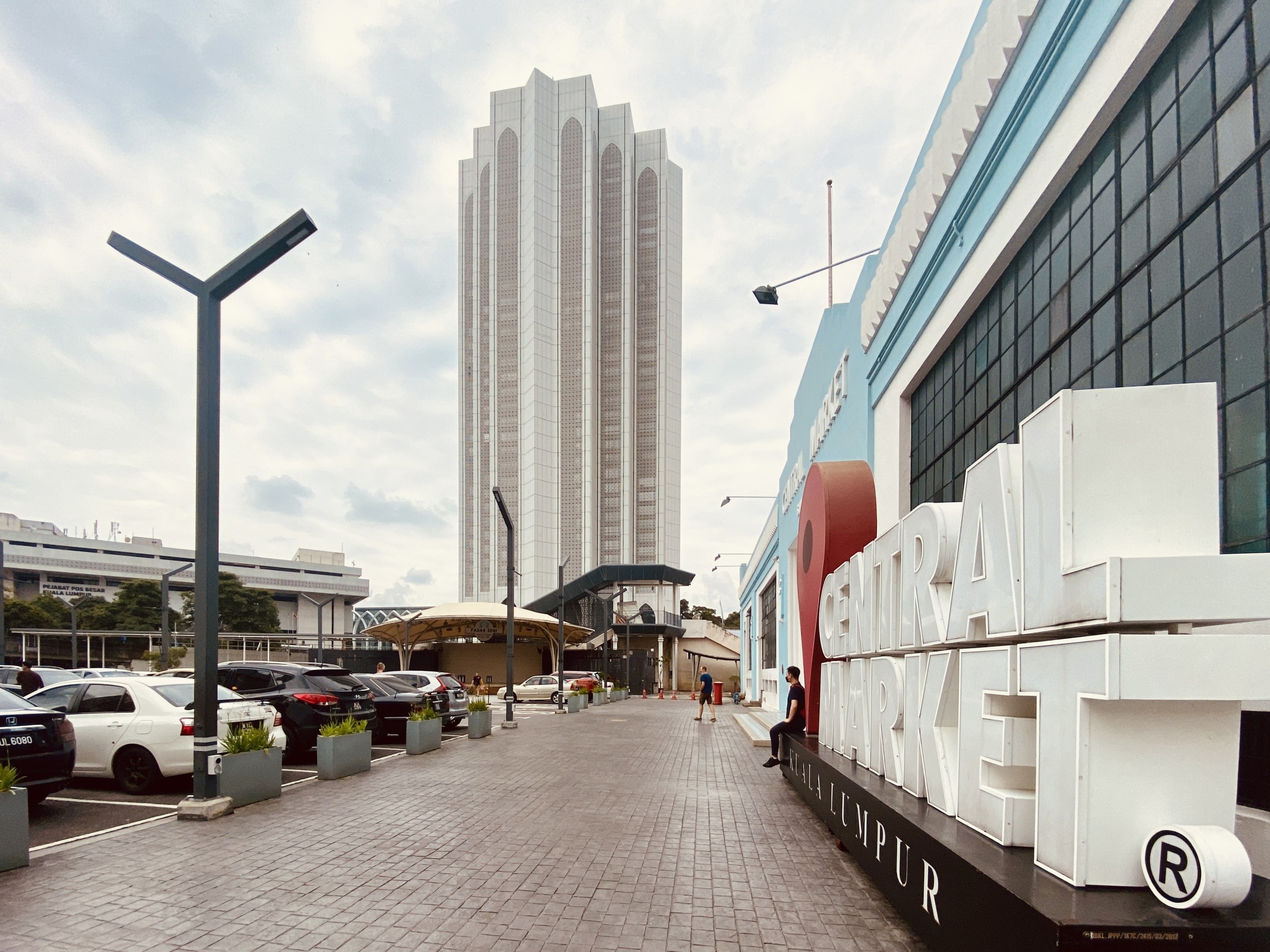 Ein belebter zentraler Markt in Kuala Lumpur, Malaysia, mit hohen Gebäuden, Straßenlaternen, Straßenschildern, Motorfahrzeugen, Fußgängern, Namensschildern, Bäumen und einem bewölkten Himmel.