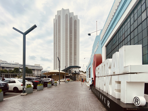 Ein belebter zentraler Markt in Kuala Lumpur, Malaysia, mit hohen Gebäuden, Straßenlaternen, Straßenschildern, Motorfahrzeugen, Fußgängern, Namensschildern, Bäumen und einem bewölkten Himmel.