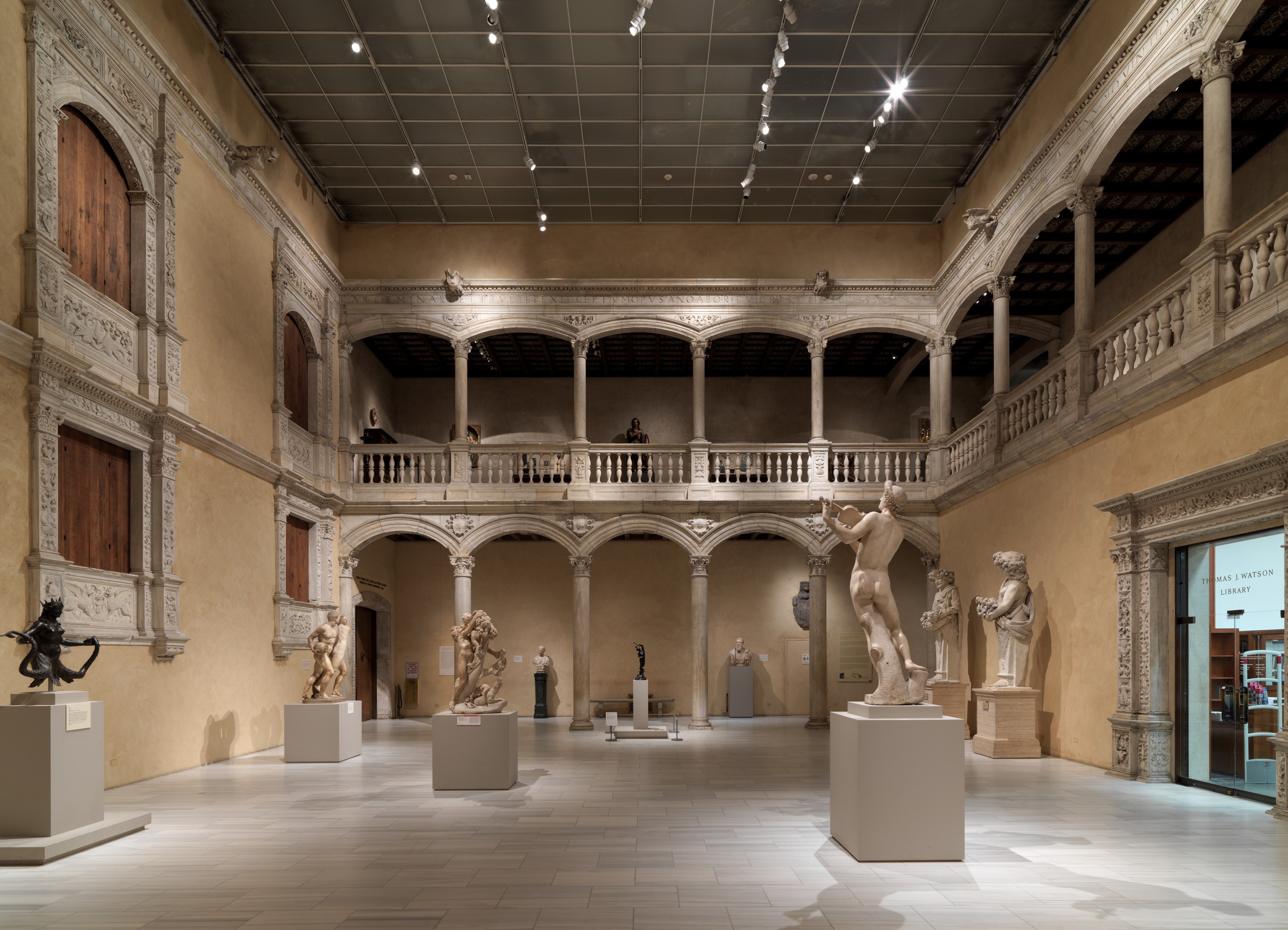 Ein geräumiger Museumsgalerie mit zahlreichen Statuen und Skulpturen auf Sockeln, mit architektonischen Elementen wie Wänden, Säulen, Geländern und einer beleuchteten Decke und einigen Besuchern, die die Ausstellungen betrachten.