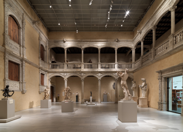 Ein geräumiger Museumsgalerie mit zahlreichen Statuen und Skulpturen auf Sockeln, mit architektonischen Elementen wie Wänden, Säulen, Geländern und einer beleuchteten Decke und einigen Besuchern, die die Ausstellungen betrachten.