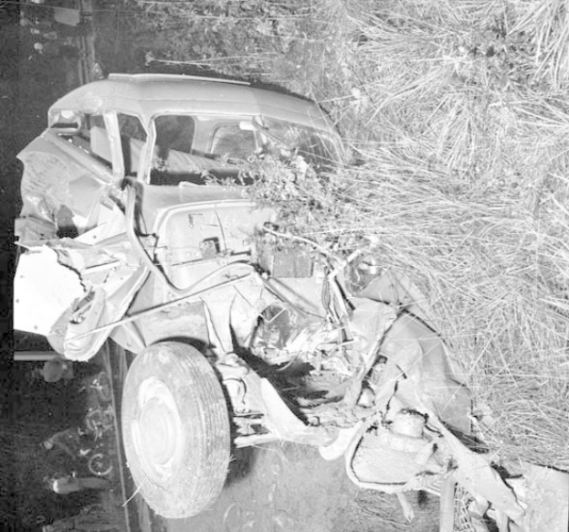 Schwarze und weiße Fotografie eines schwer beschädigten Autos mit eingedellter Front und zersplitterten Fenstern am Straßenrand, mit einem Baum und verstreuten Gegenständen im Hintergrund.