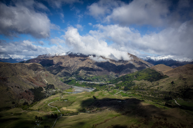 Ein atemberaubender Blick von der Spitze eines Berges in Queenstown, Neuseeland, mit saftig grünem Gras, Bäumen, einer kurvenreichen Straße und einem Himmel voller weißer, flauschiger Wolken.