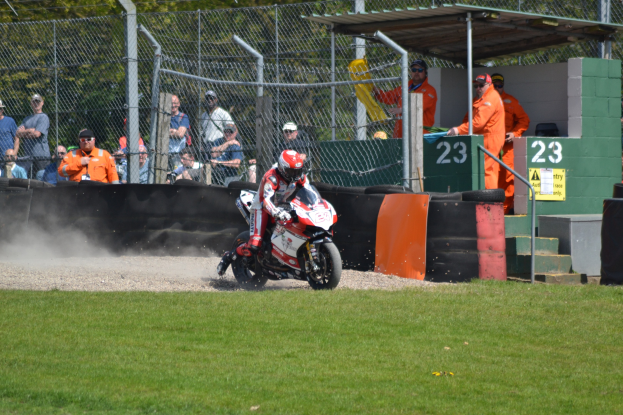 Eine Person in Helm und Handschuhen fährt auf einem grasbewachsenen Feld ein Motorrad, mit Zuschauern im Hintergrund in Mützen und Schutzbrillen, Maschendrahtzaun, Treppen mit Geländern und einem nummerierten Schuppen mit Texttafeln.