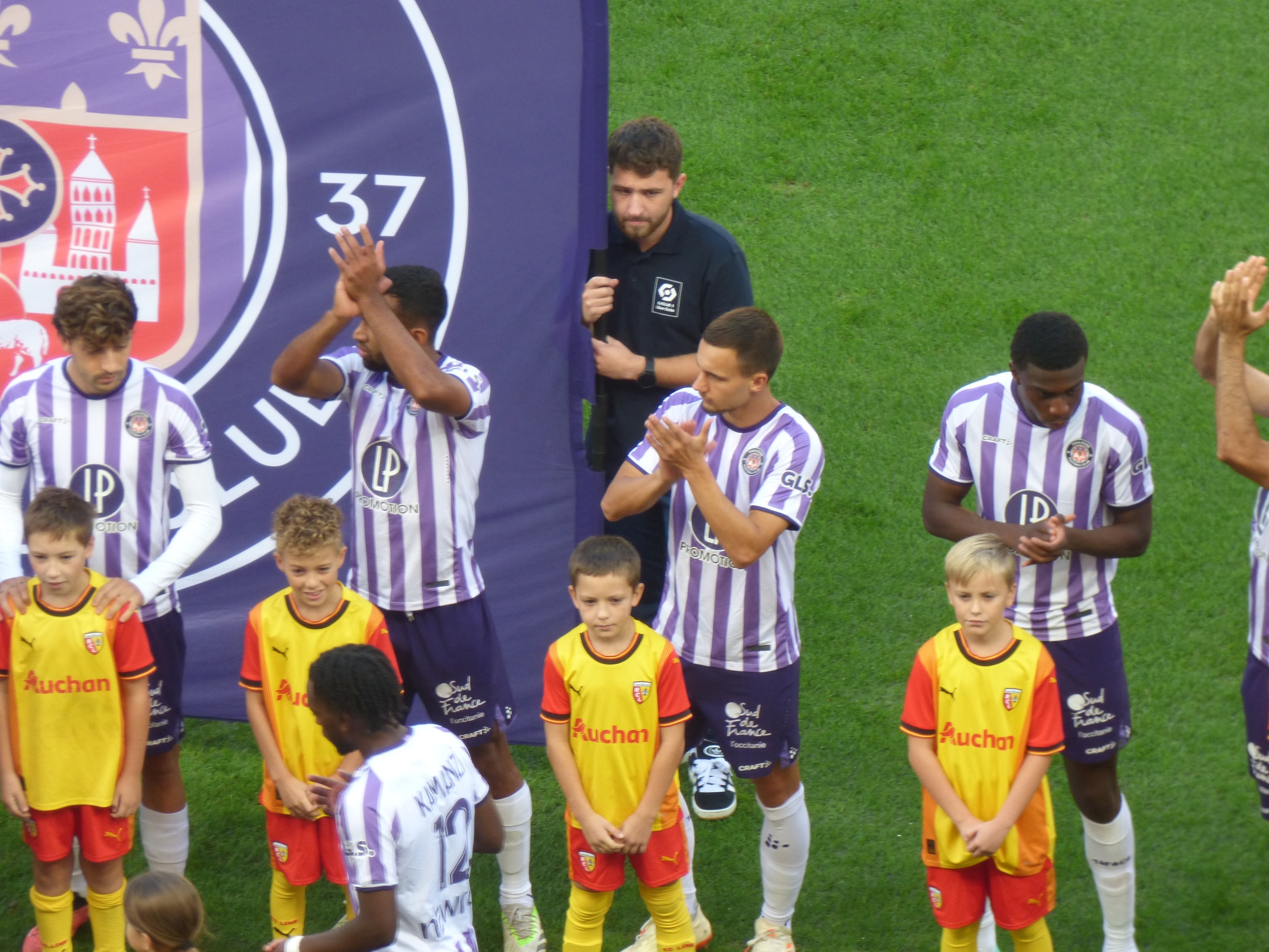 Gruppe junger Jungs auf einem Fußballfeld, die klatschen, mit einem Banner und Logos im Hintergrund auf grünem Rasen.