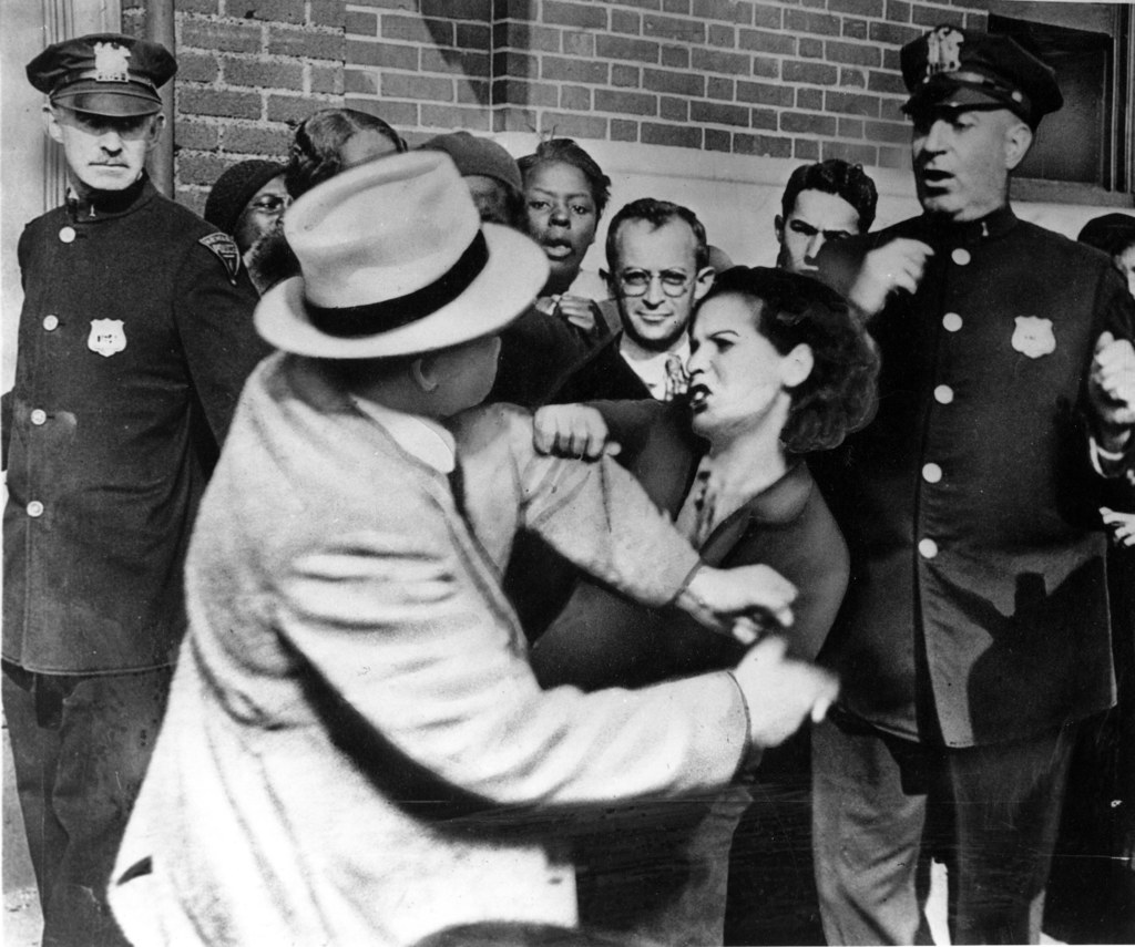 Zwei Personen kämpfen auf einem Schwarz-Weiß-Bild, mit mehreren anderen im Hintergrund, einige tragen Mützen und eine Wand im Hintergrund.