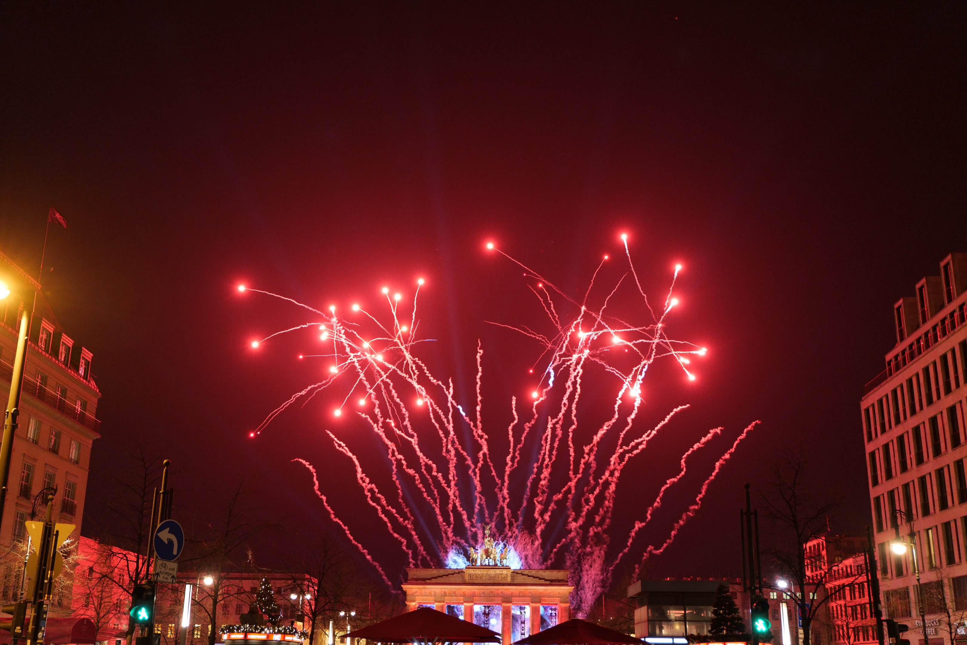 Eine belebte Stadtstraße an einem Silvesterabend in Berlin, mit Gebäuden, Bäumen, Laternenpfählen, Verkehrsampeln, Schildern, Zelten, Menschen und einem prächtigen Feuerwerk am Himmel.