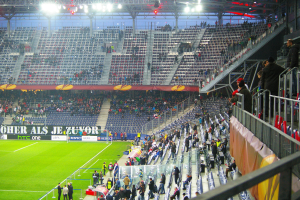 Ein volles Stadion mit Zuschauern, die ein Fussballspiel verfolgen, mit einigen Personen, die auf dem Feld stehen oder sitzen, einem Geländer und einer Fahne auf der rechten Seite und einem leeren Stadion mit Deckenlampen im Hintergrund.