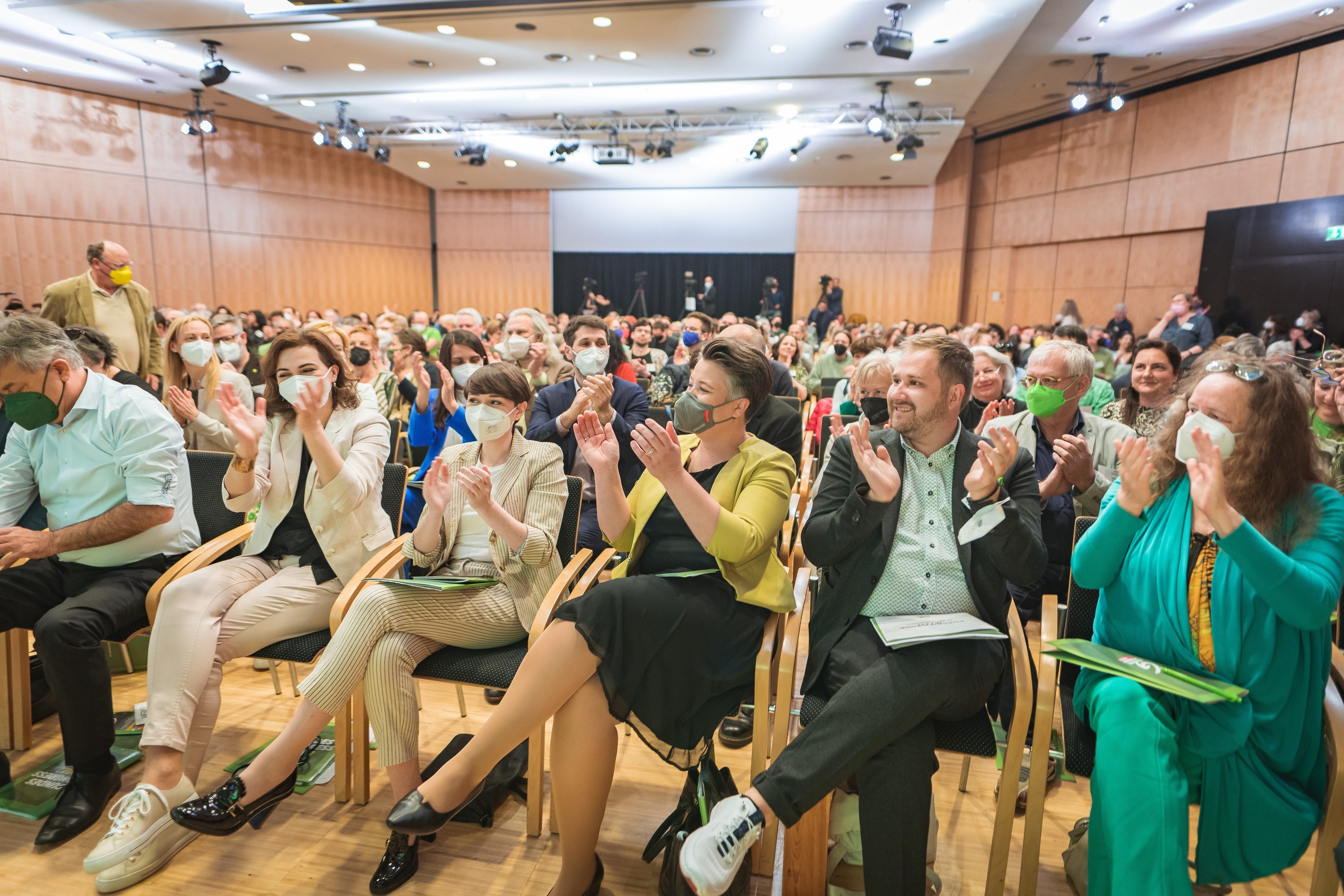 Eine Gruppe von Menschen in Stühlen, die vor einem Publikum klatschen, einige tragen Masken, mit Taschen auf dem Boden, in einem Konferenzsetting mit einem Bildschirm und Deckenleuchten.