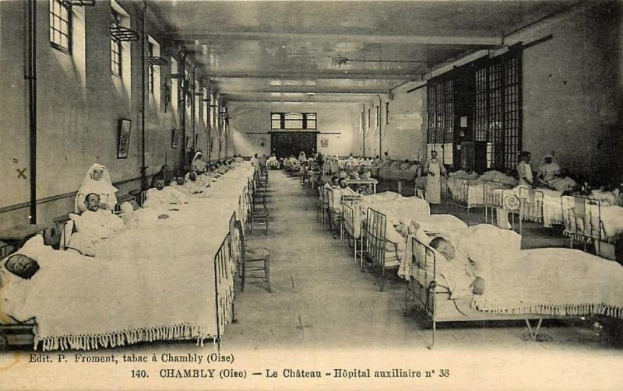 Schwarzes und weißes Foto einer Krankenhausstation mit mehreren Betten, Patienten, Möbeln, Wandrahmen, Deckenlampen und Text am unteren Rand.