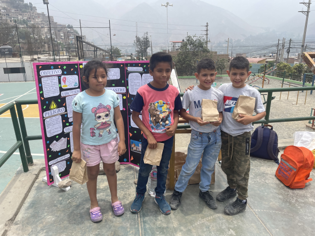 Gruppe von Kindern auf einem Basketballfeld mit Papiertüten in den Händen, Tüten und Gegenstände rechts und eine Tafel mit Text links, vor einer Außenkulisse mit Zaun, Pfosten und Gebäuden.