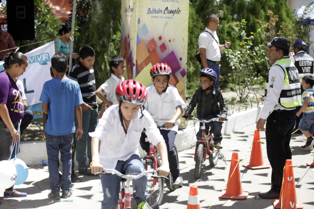 Eine Gruppe von Kindern, die auf Fahrrädern eine Straße entlangfahren, mit Verkehrskegeln, einige tragen Helme, andere stehen daneben, mit einer Fahne, Bäumen und Gebäuden im Hintergrund.
