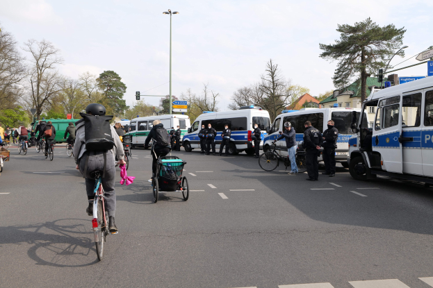Eine Gruppe von Menschen, die auf Fahrrädern eine Straße entlangfahren, mit Polizeiwagen, einige tragen Helme und haben Taschen oder schieben Kinderwagen, gesäumt von Bäumen, Gebäuden und Verkehrszeichen unter einem bewölkten Himmel.