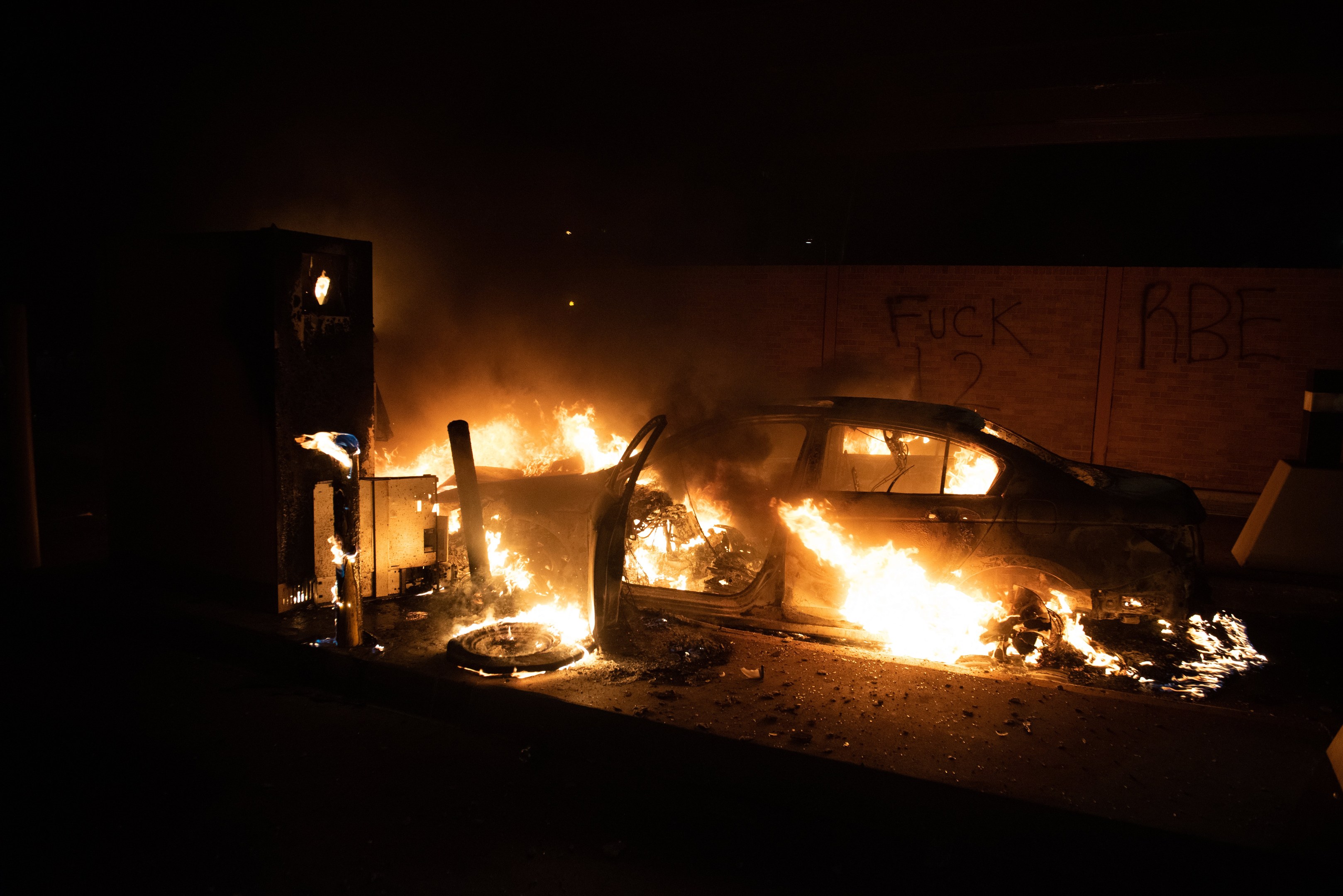 Ein brennendes Auto am Straßenrand bei Nacht, mit einer beleuchteten Wand mit Graffiti im Hintergrund.