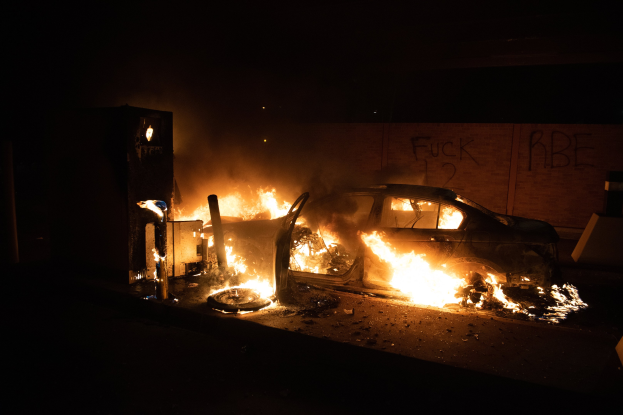 Ein brennendes Auto am Straßenrand bei Nacht, mit einer beleuchteten Wand mit Graffiti im Hintergrund.