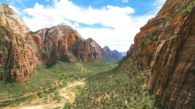 Atemberaubender Ausblick auf den Zion-Nationalpark in Utah mit saftig grünen Bäumen, sanften Hügeln, einem friedlichen Gewässer im Vordergrund und einem Himmel mit flauschigen weißen Wolken.