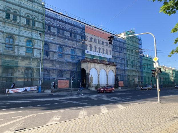 Großes Gebäude mit Gerüst, das renoviert wird, umgeben von Fahrzeugen, Passanten, Ampeln, Schildern, Straßeninfrastruktur, Bäumen und einem klaren blauen Himmel.