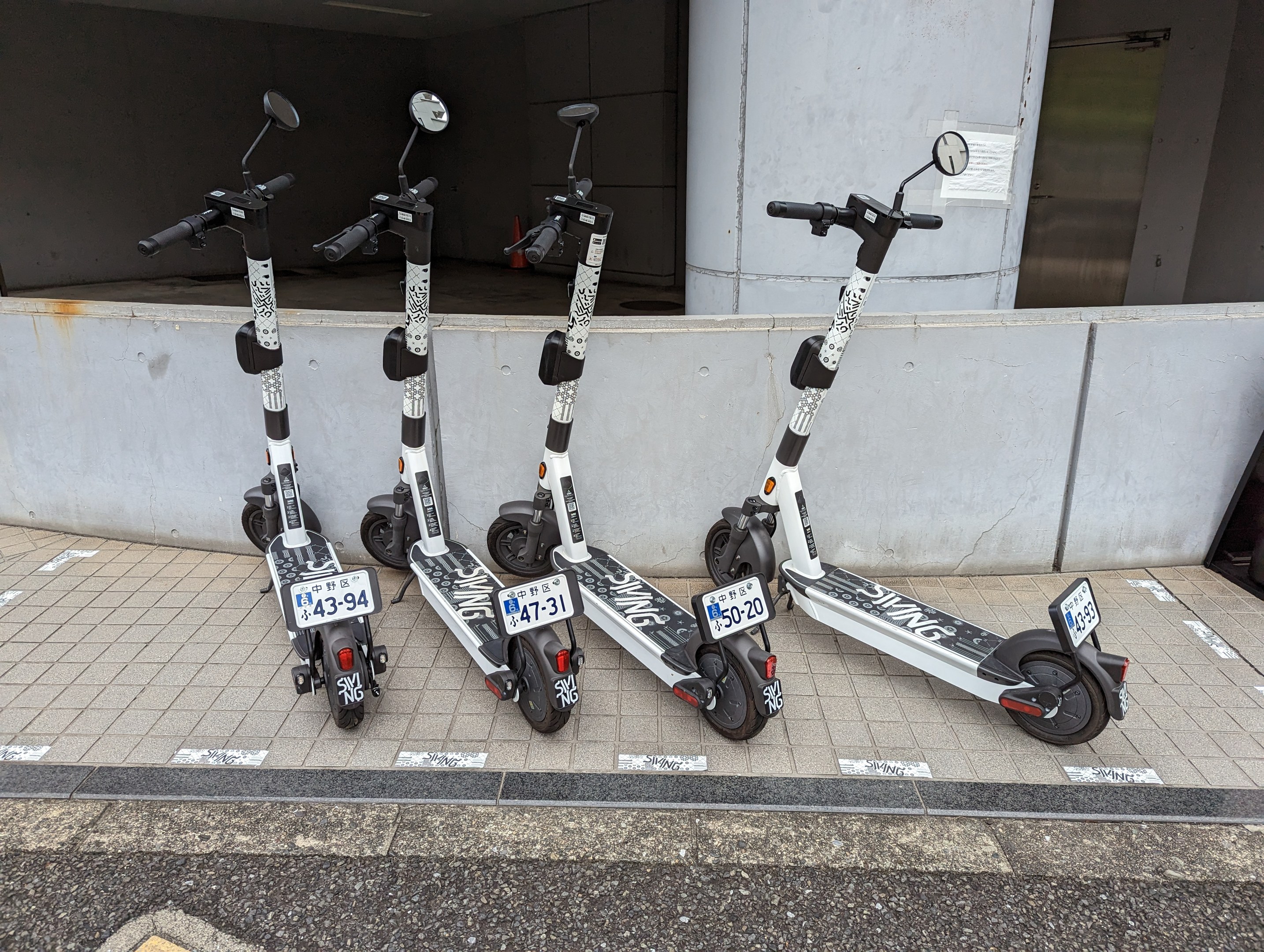 Drei schwarze und weiße E-Scooter mit Nummern und Text stehen nebeneinander auf einem Gehweg vor einem Gebäude mit einer Wand und einer Glastür im Hintergrund.