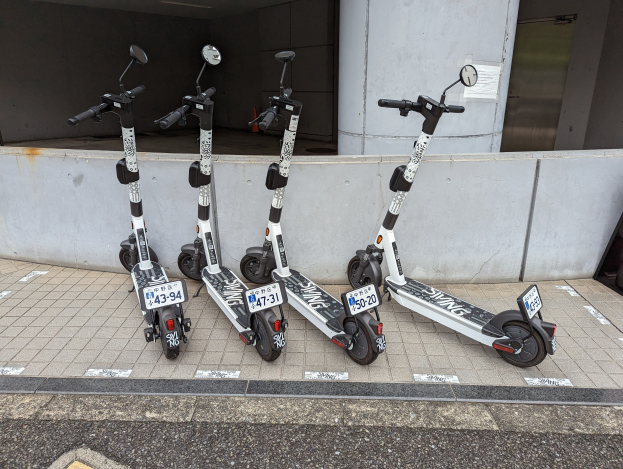 Drei schwarze und weiße E-Scooter mit Nummern und Text stehen nebeneinander auf einem Gehweg vor einem Gebäude mit einer Wand und einer Glastür im Hintergrund.