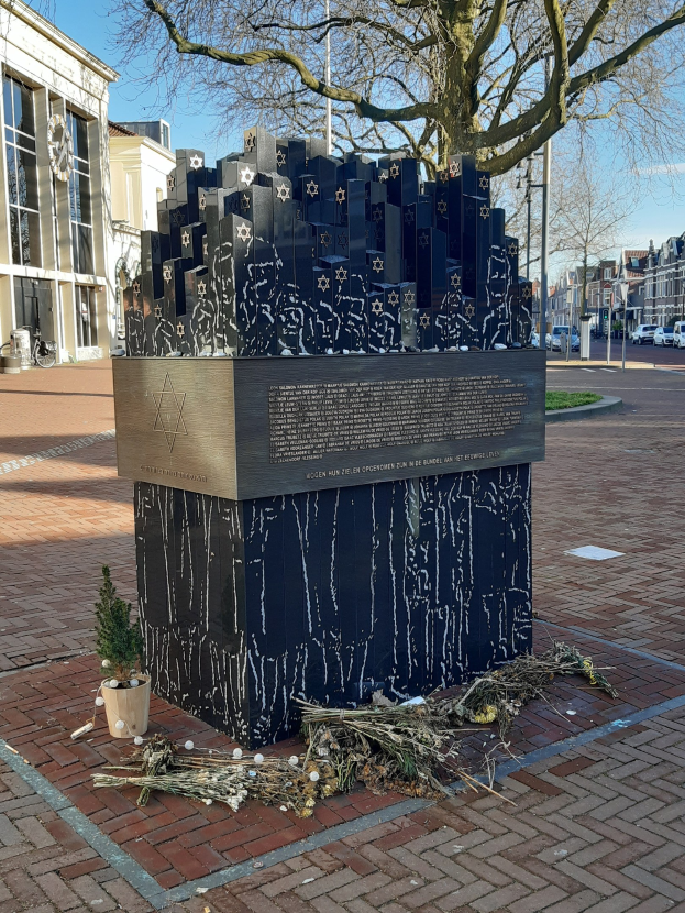 Denkmal in einem Stadtplatz gewidmet an Holocaust-Opfer, mit einem Baum davor, einem Topfgewächshaus auf dem Boden, verstreuten trockenen Blättern und Gebäuden, Fahrzeugen, Masten und Bäumen im Hintergrund.