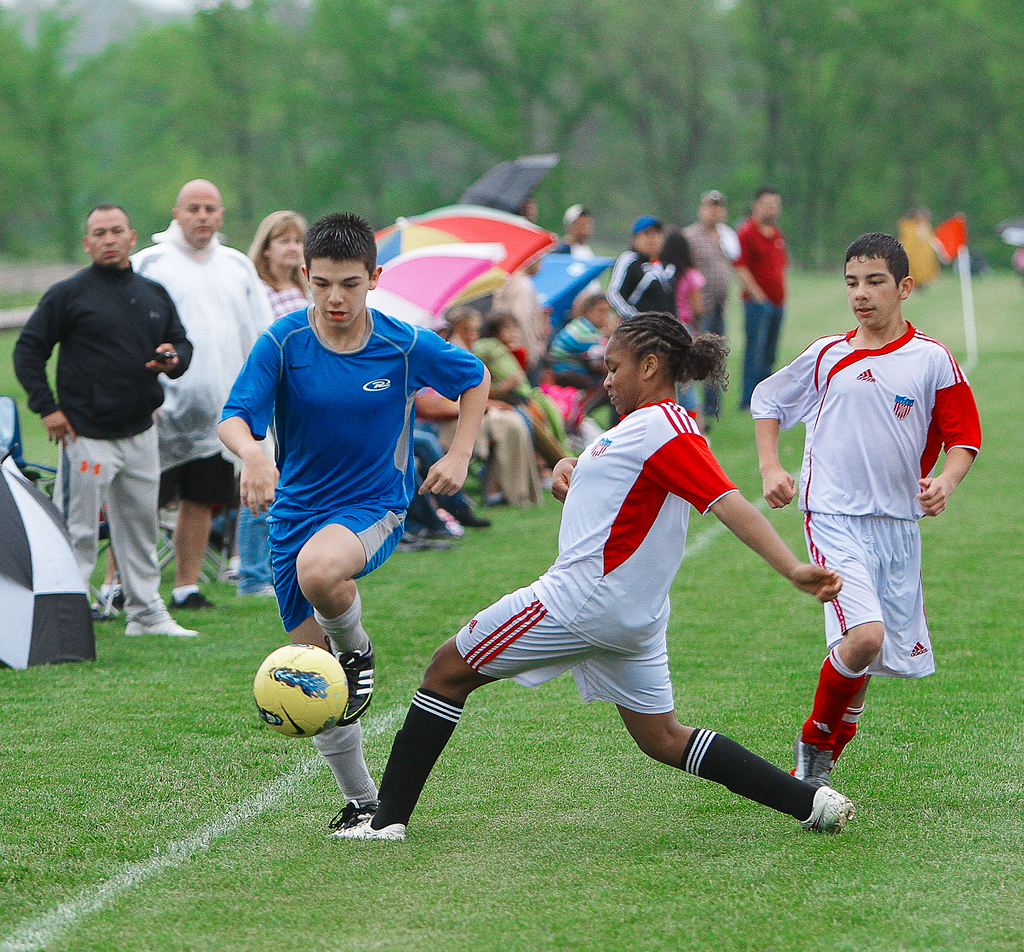 Drei Personen spielen Fussball mit einem gelben Ball, während eine Gruppe von Menschen hinter ihnen steht und sitzt und Regenschirme hält, mit Bäumen im Hintergrund.