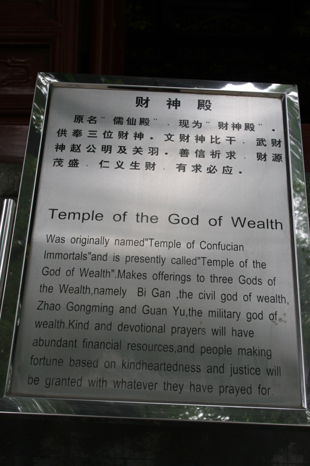 Ein Tempel, der dem Gott des Reichtums gewidmet ist, mit einer Namensschild im Vordergrund und einer Tür im Hintergrund, umgeben von üppiger Vegetation.