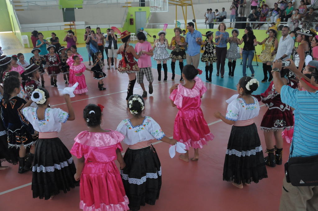 Eine Gruppe von Kindern in Kostümen tanzt in der Mitte des Bildes, während das Publikum von der oberen rechten Seite zusieht.