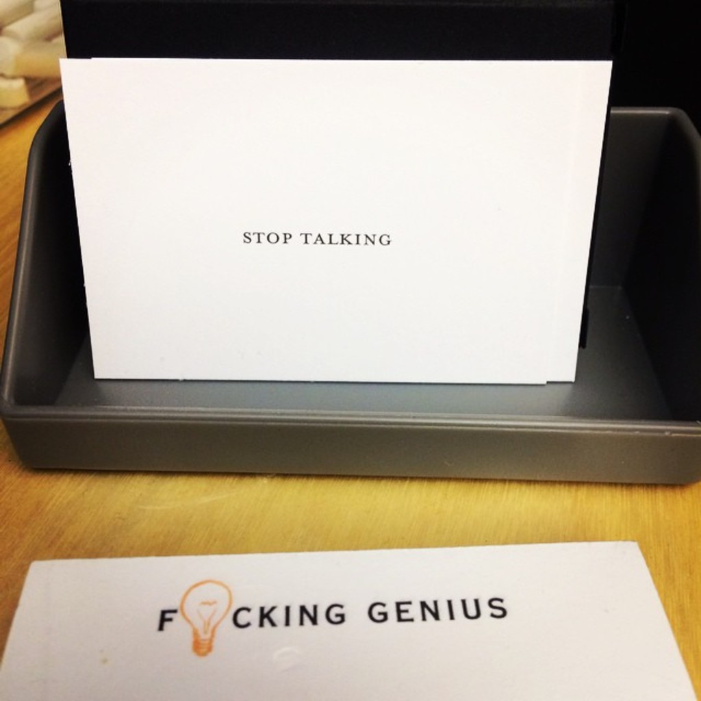 Ein Druckerbox mit einem Blatt Papier darauf, das den Text "Aufhören zu sprechen" anzeigt, und einem weiteren Blatt auf dem Tisch, das den Text "Fucking genius" anzeigt.