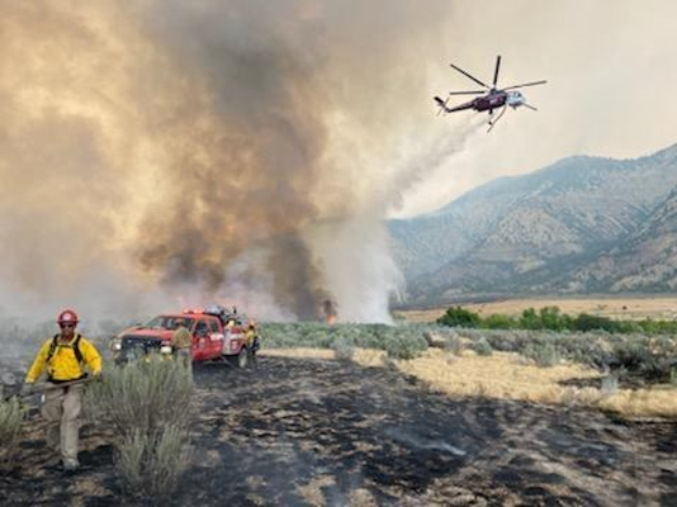 Ein Hubschrauber überfliegt ein Feld mit einem Feuerwehrauto im Hintergrund, der Himmel ist von Rauch erfüllt und Hügel sind in der Ferne zu sehen, mit einigen Menschen, Fahrzeugen und Pflanzen auf dem Gebiet verteilt.