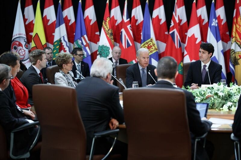 Gruppe von Menschen, die um einen Tisch mit Flaggen im Hintergrund sitzen und diskutieren, mit Mikrofonen, Gläsern, Blumen, Papieren und anderen Gegenständen auf dem Tisch, wahrscheinlich im Zusammenhang mit einem Kanada-Kanada-Handelsabkommen.